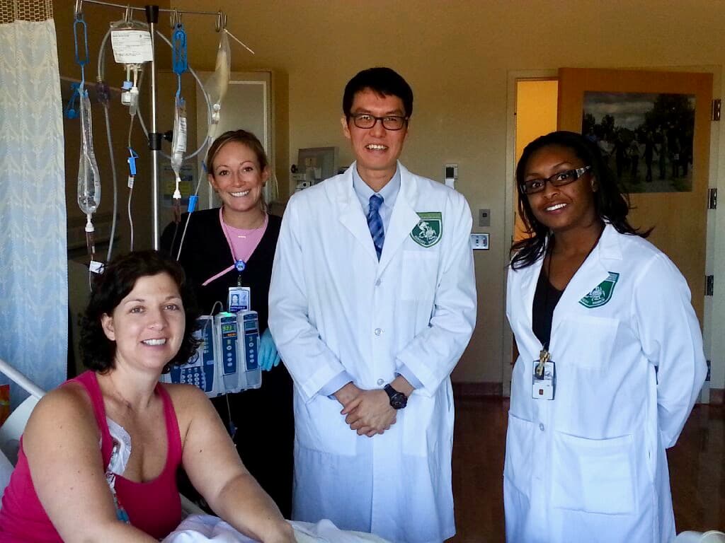 Melinda Bachini on her hospital bed, with 2 doctors and a nurse standing next to her.